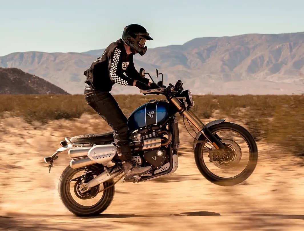 Triumph Scrambler 1200 XE scrambling across sandy plains performing a wheelie. Rider wearing limited edition Scrambler 1200 clothing